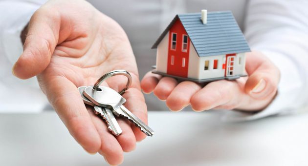 A person holds a key in one hand and a small house model in the other, symbolizing home ownership and new beginnings.